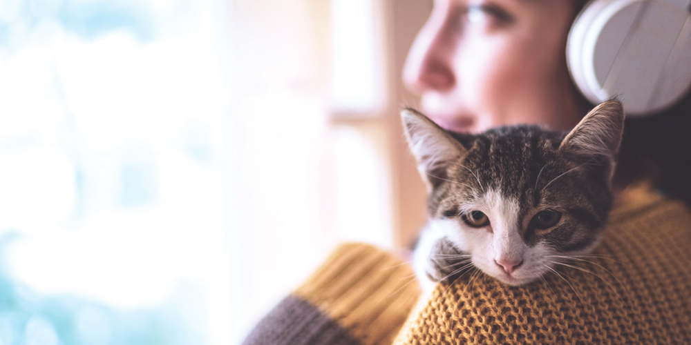 Woman with headphones holding cat while looking out a window